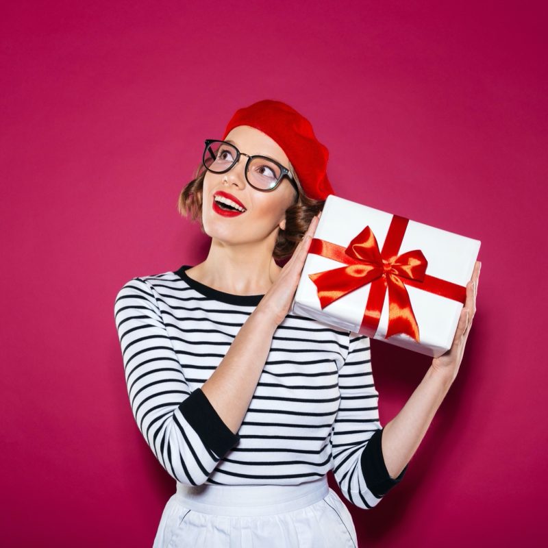 Intrigued happy ginger woman in eyeglasses holding gift box near the ear and looking up over pink background