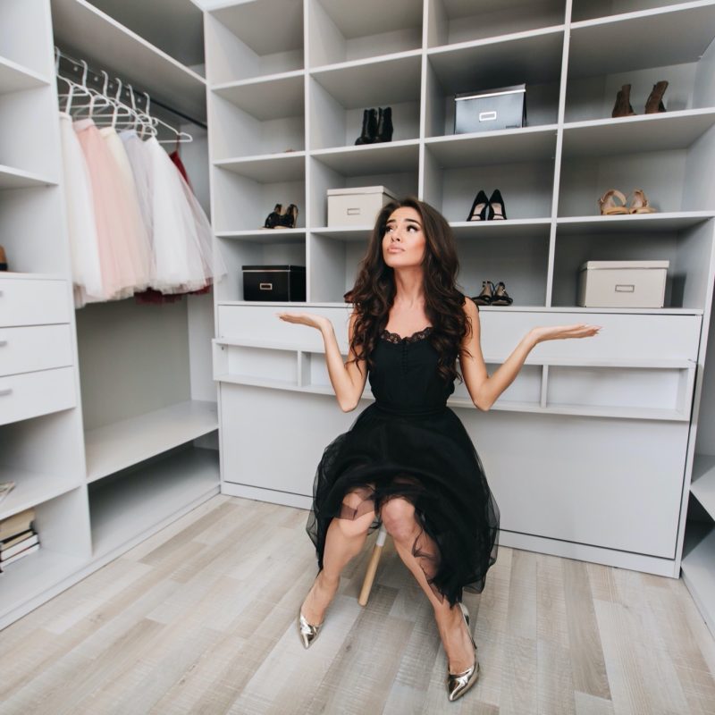 Young brunette woman sitting in a huge dressing room thinks over the choice of clothes, she is dressed stylish black outfit and silver shoes, expressing true positive face emotions.